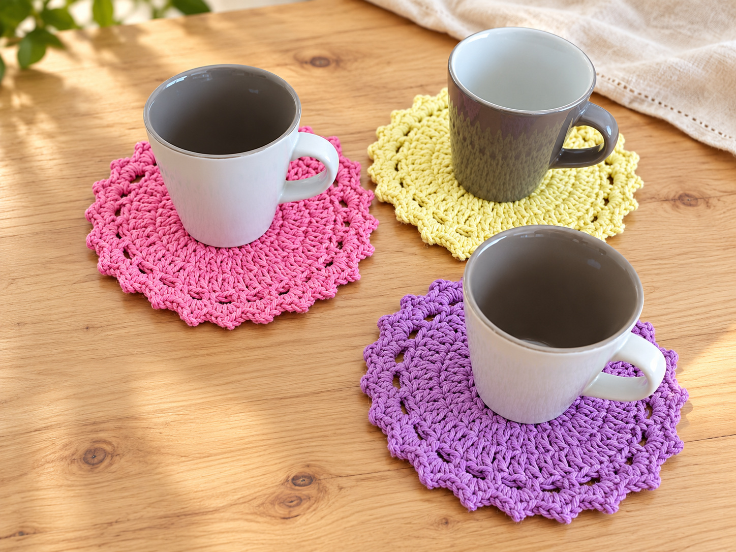 Gray mugs on pink, pale yellow, and purple scalloped crochet coasters on a wooden table.