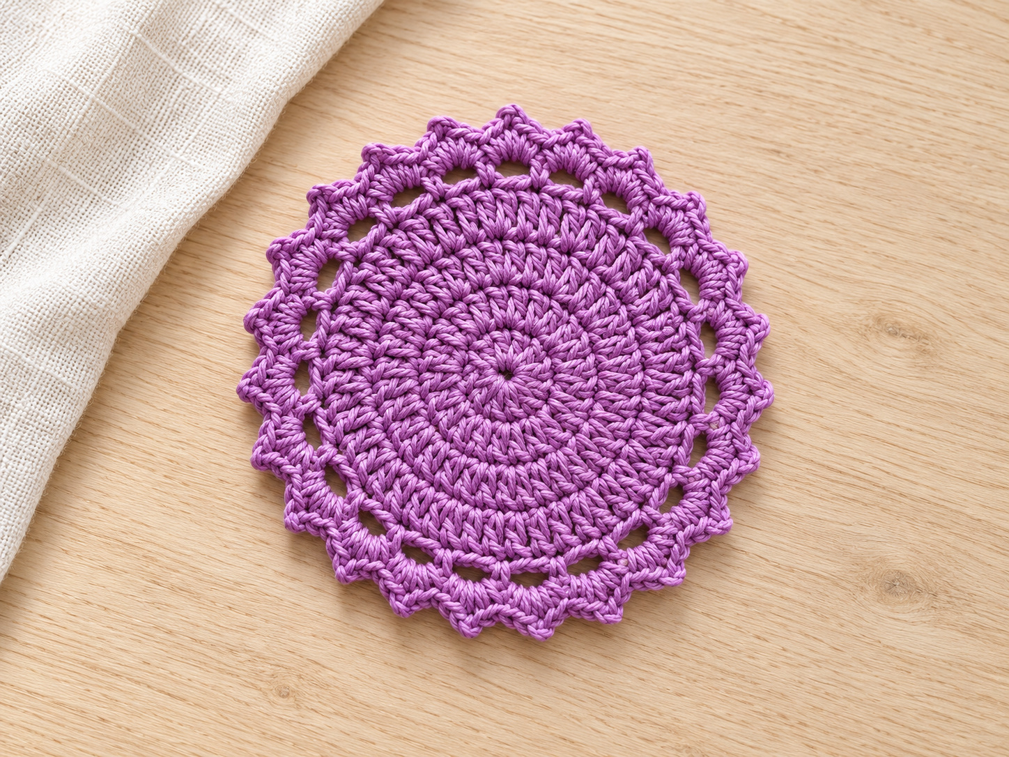 A purple round crochet coaster with a solid center, open chain spaces, and scalloped edging beside ivory linen.