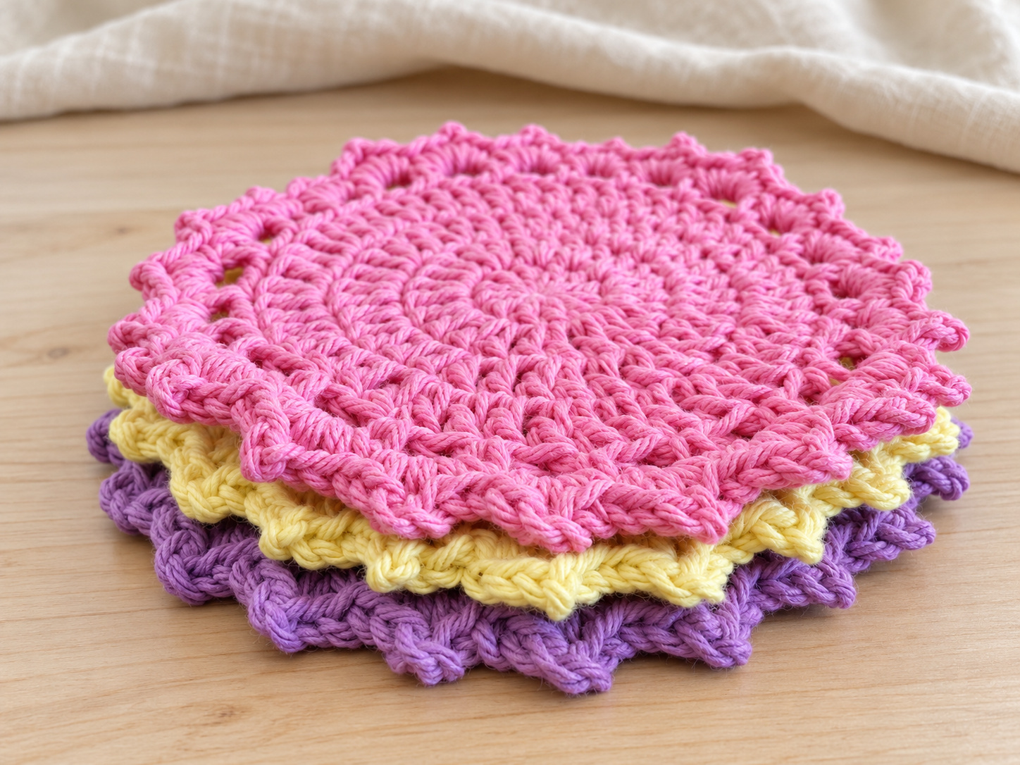 Three scalloped crochet coasters stacked in pink, pale yellow, and purple on a pale wooden table.