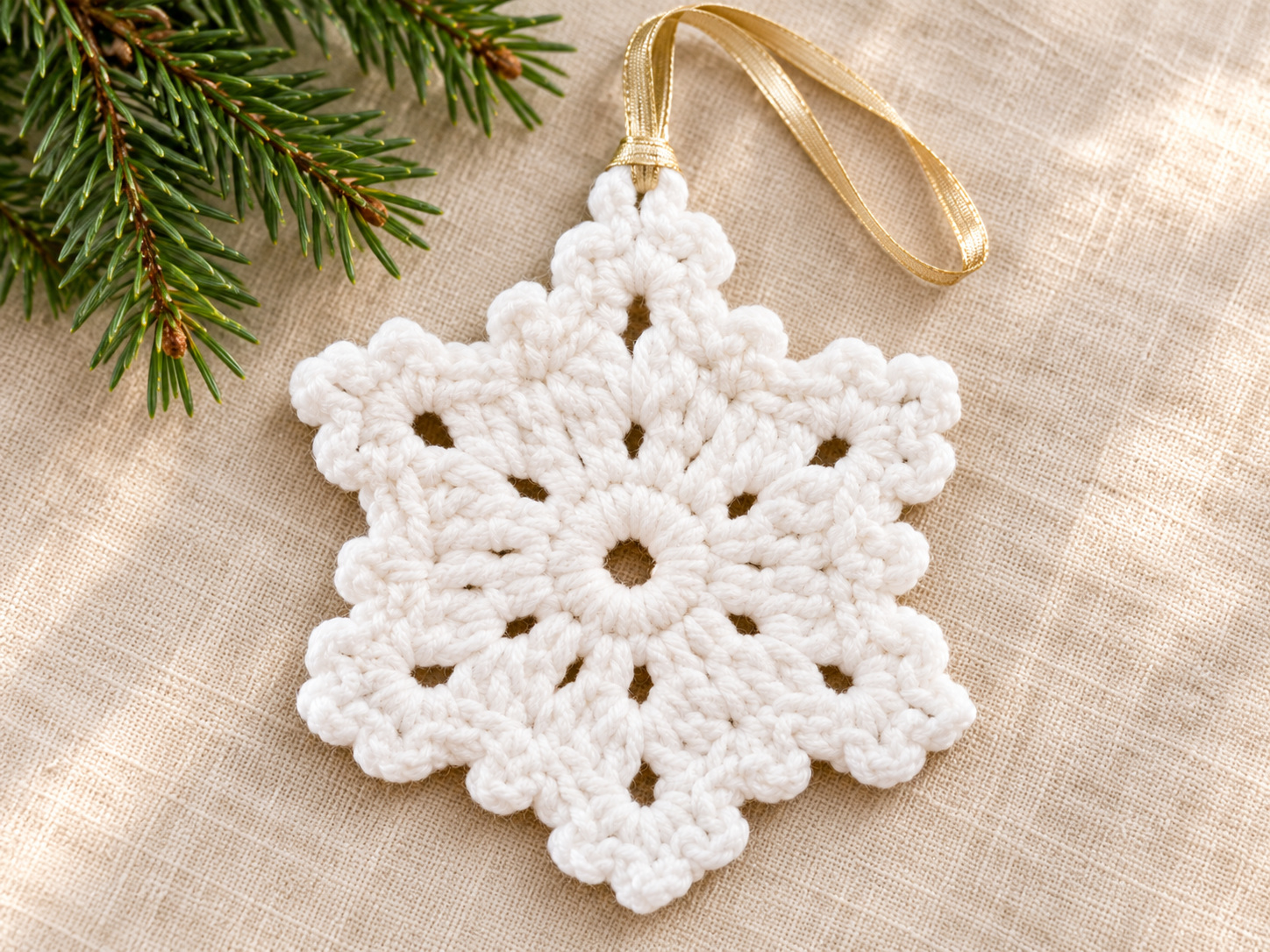 A white crochet snowflake with a gold hanging ribbon on ivory linen beside an evergreen sprig.